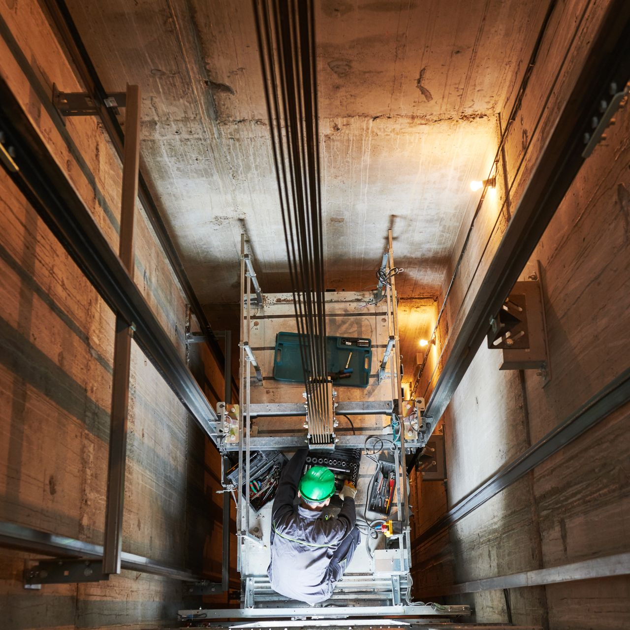 Intérieur d'une cage d'ascenseur avec un ouvrier portant un casque, inspectant les machines.