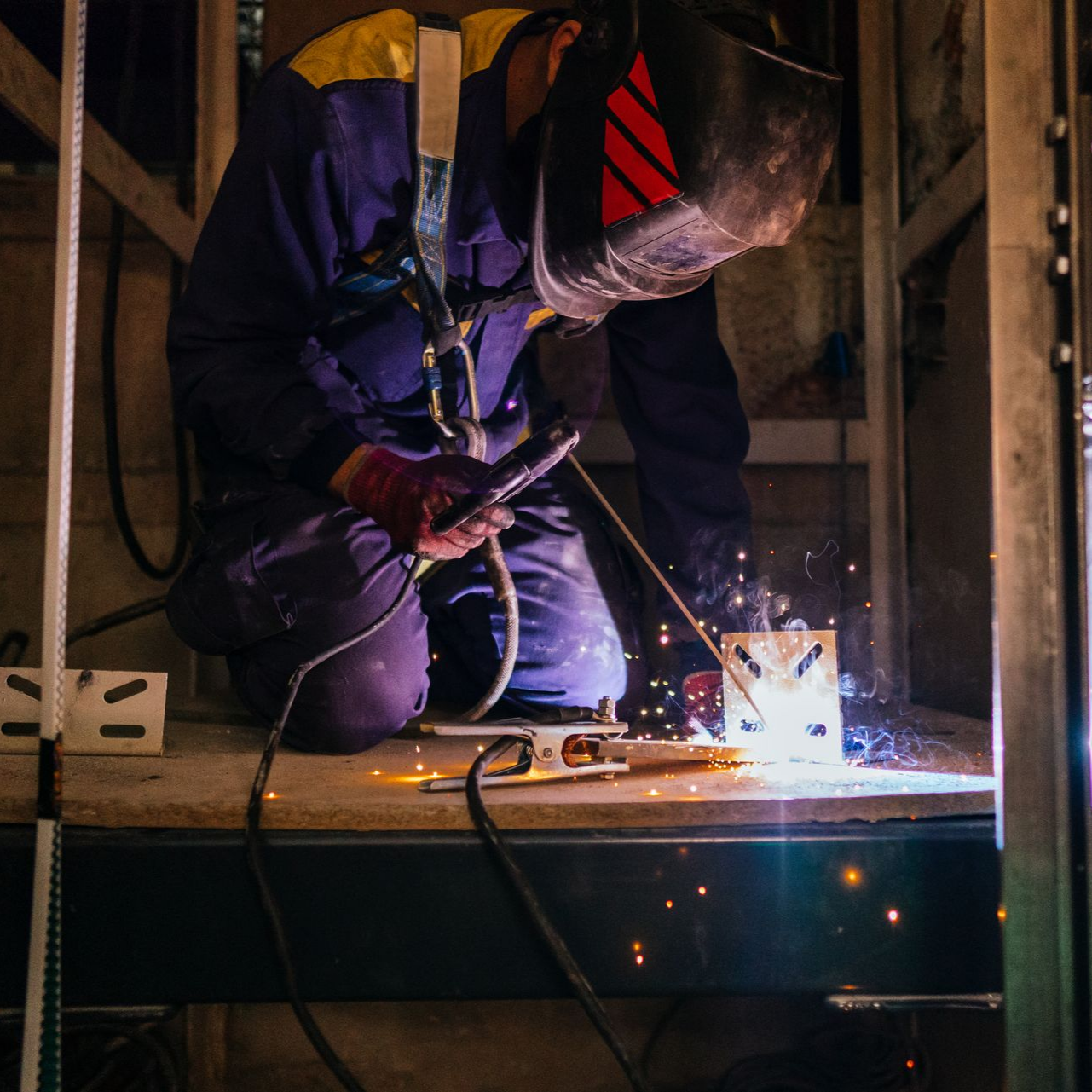 Soudeur portant un masque soudant du métal, agenouillé sur une plate-forme métallique.