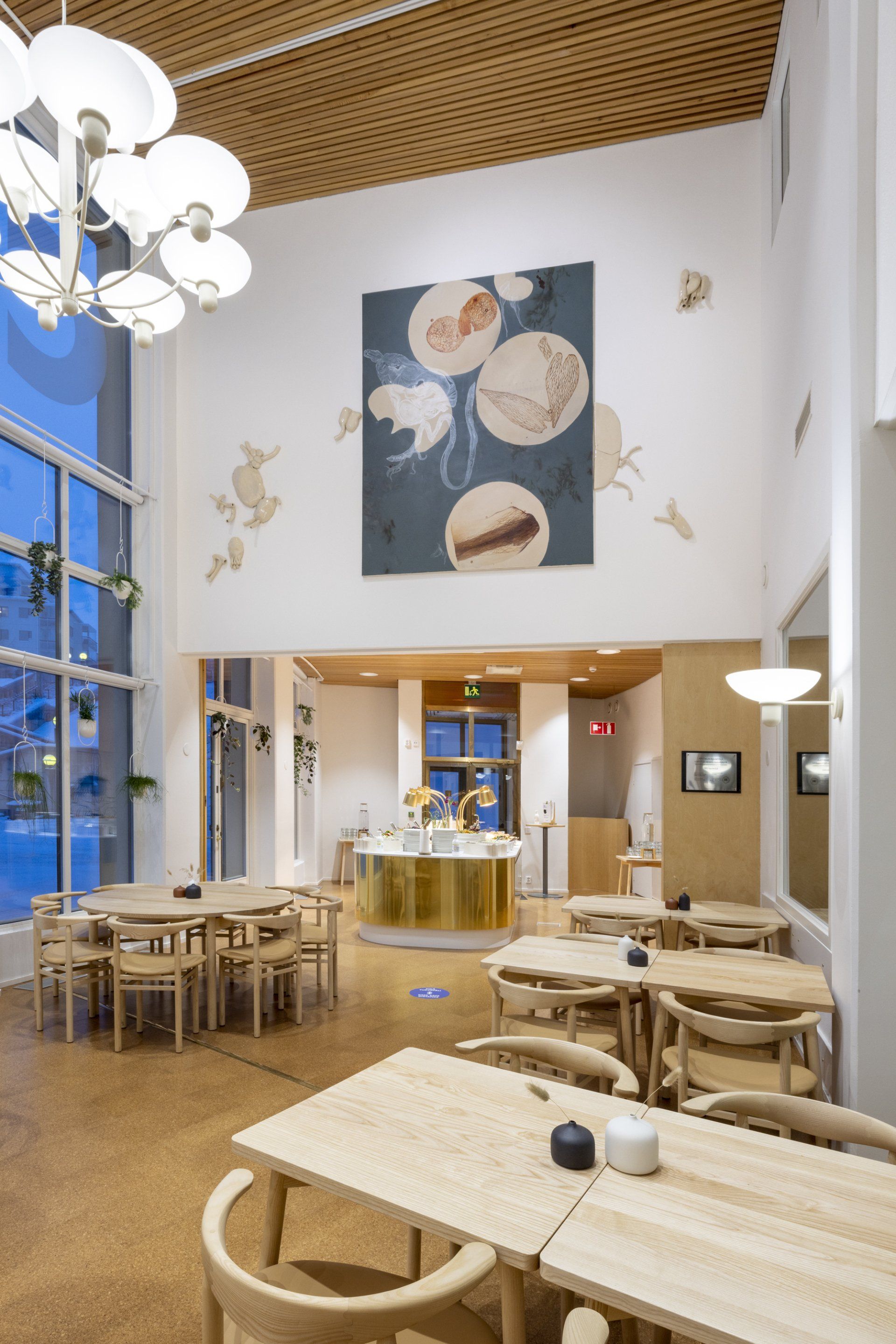 Ravintolasuunnitelma. Puiset kalusteet, messinkinen tiski ja taideteos seinällä. Ikkunalla roikkuvat kasvit ruukuissa. 