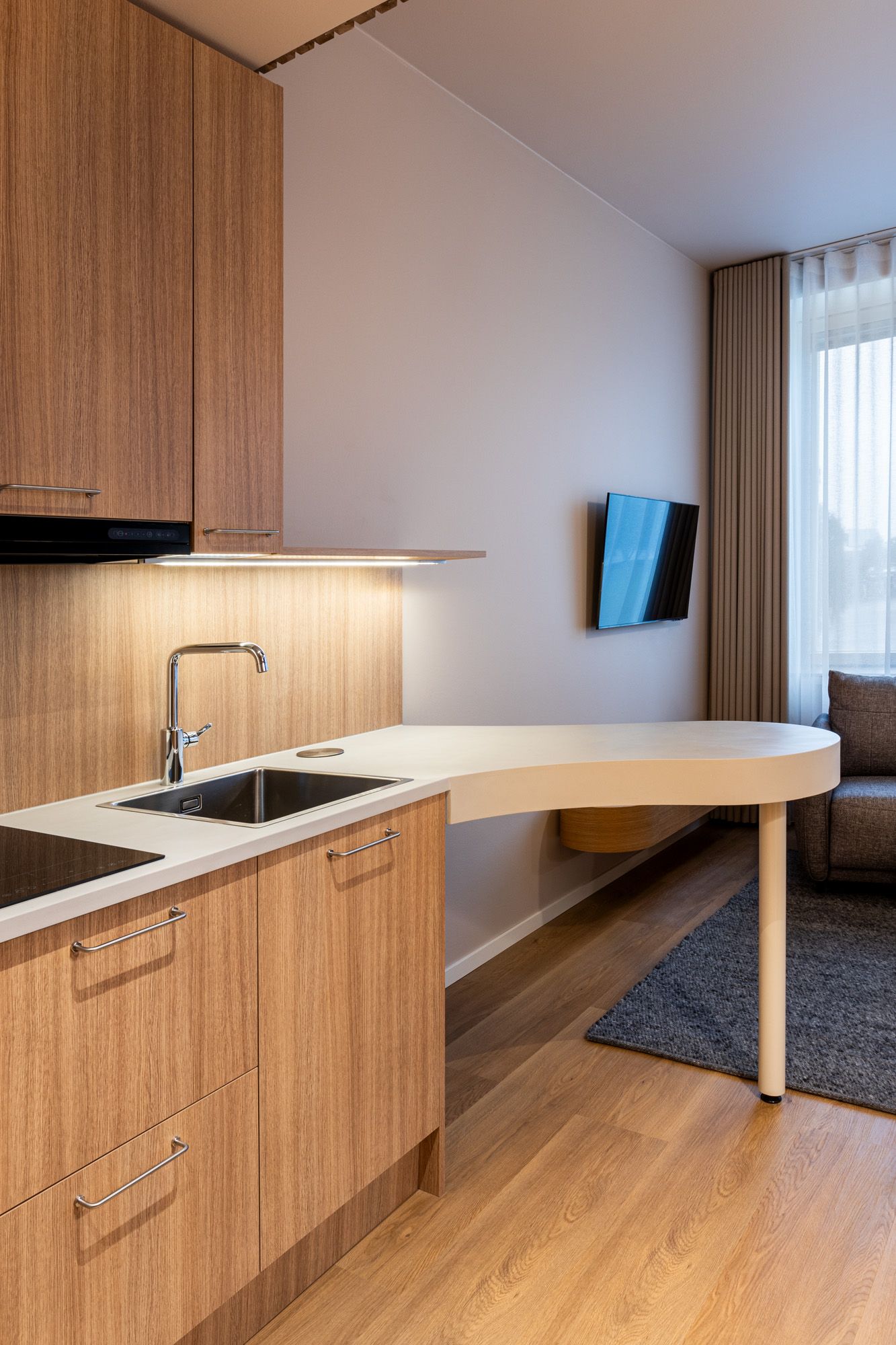 Hotellihuoneen pähkinäpuinen minikeittiö. Keittiöön integroitu veistoksellinen ruokapöytätaso. 