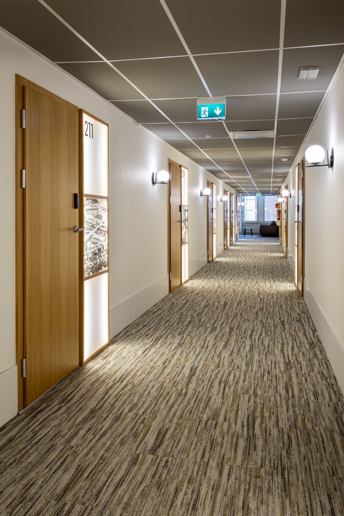 Hotellisuunnittelu - tammi ovet hotellikäytävässä. Ovien vieressä valopaneelit puukehillä. 