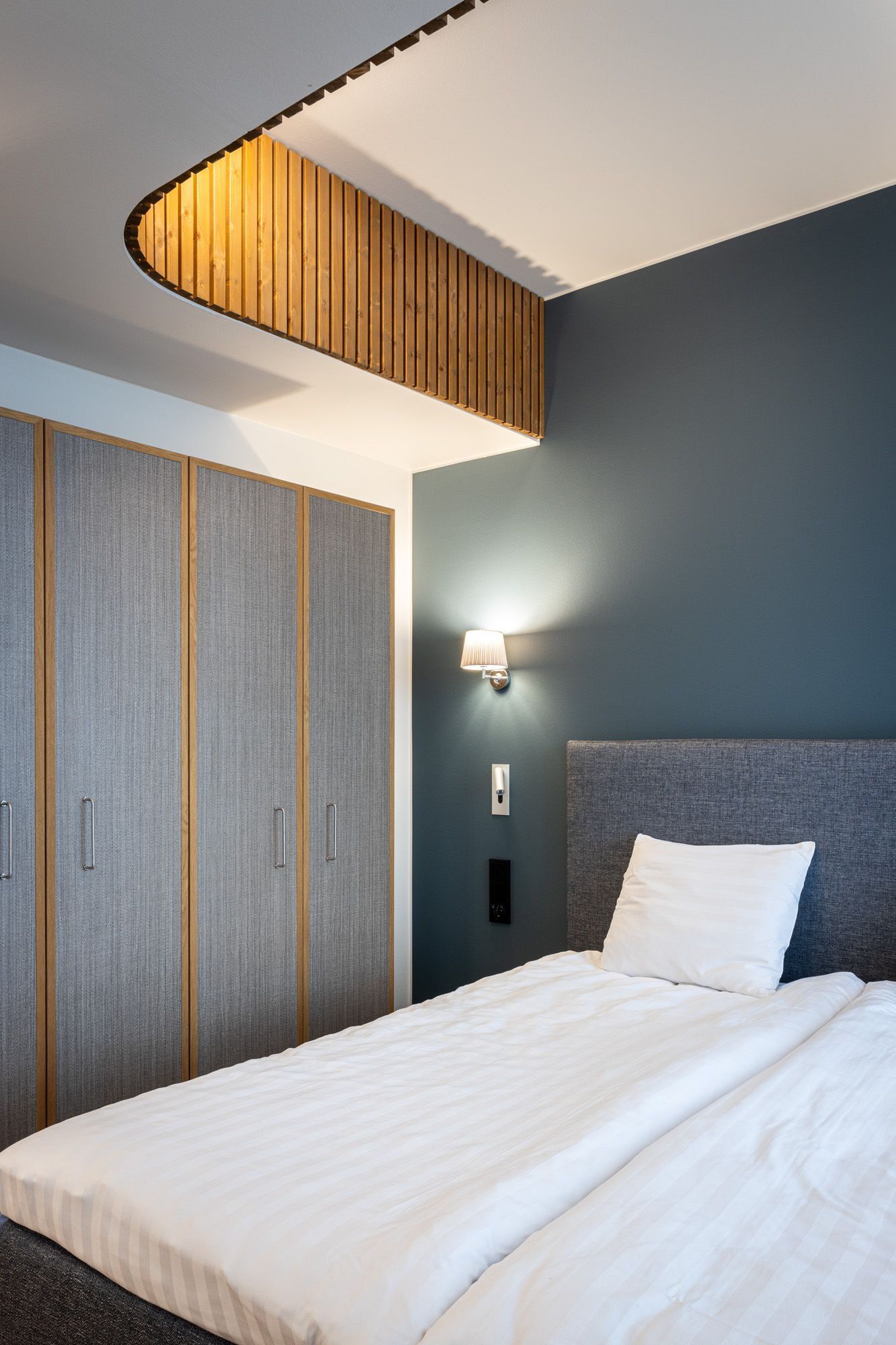 Hotellihuoneen harmaa sängynpääty ja vaatesäilytyskaluste. Takana sininen seinä. 