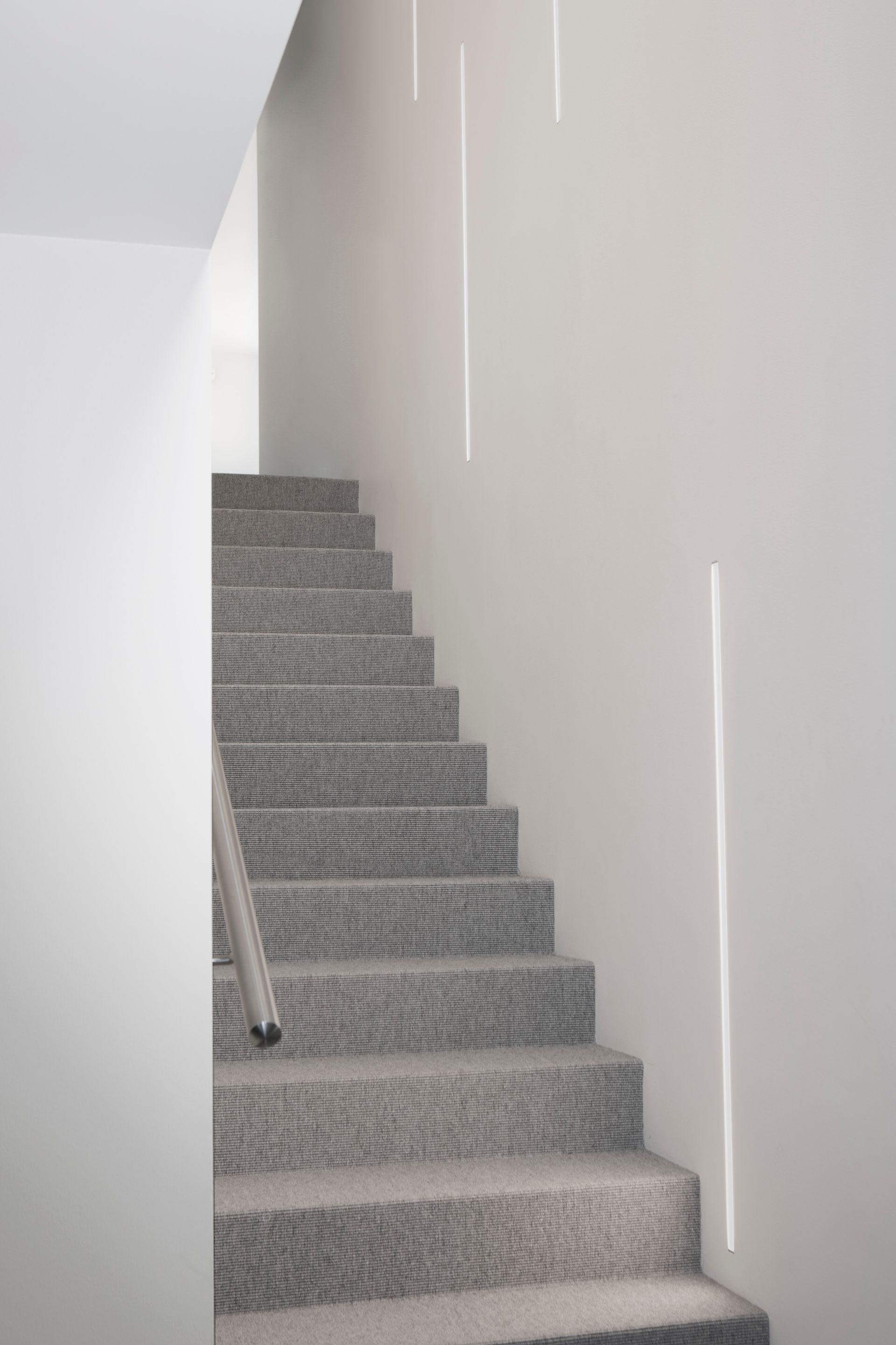 Omakotitalon portaikon suunnitelma. Porrasaskelmissa harmaa kokolattiamatto. Seinässä upotetut linjavalaisimet. 
