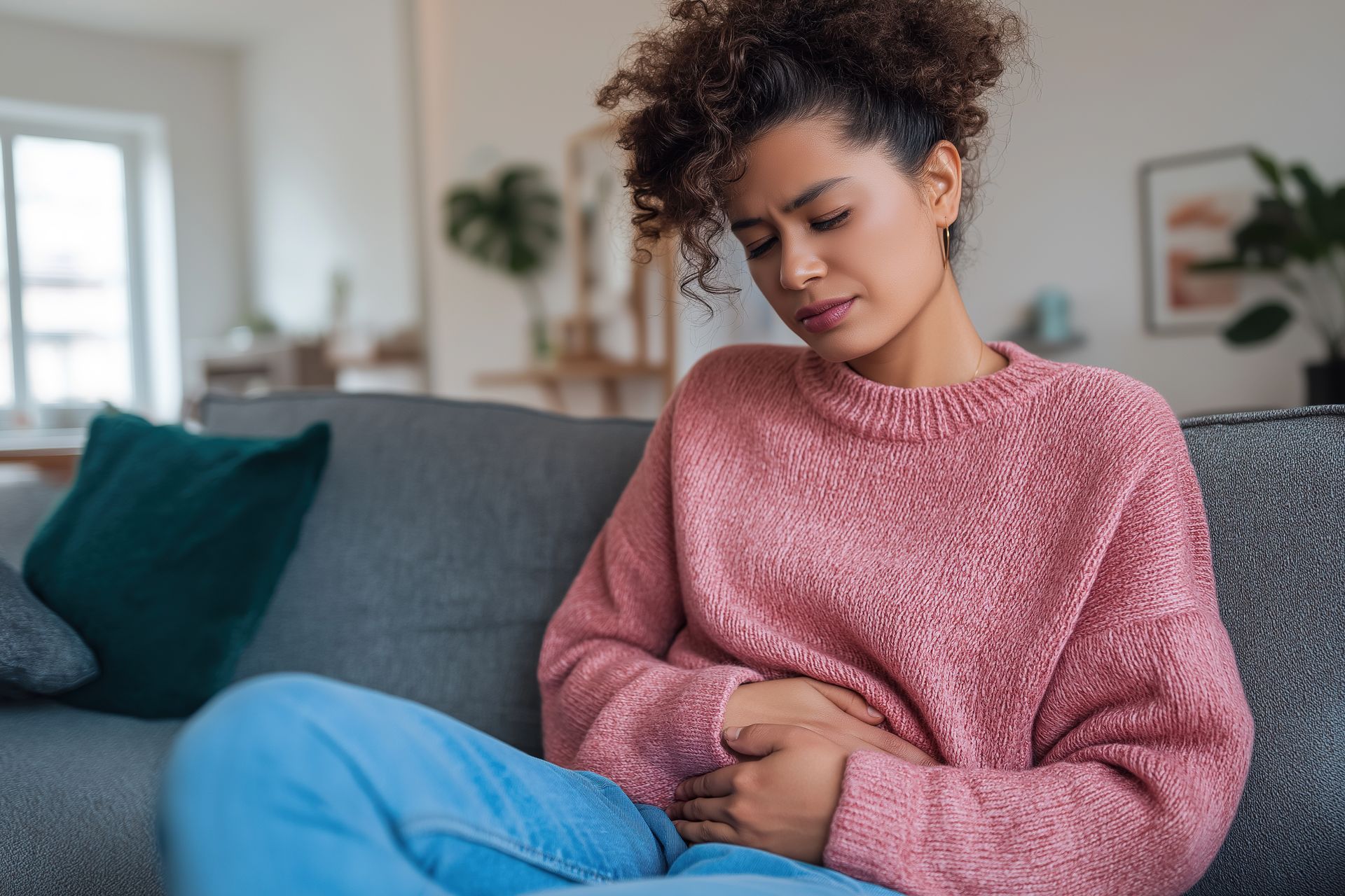 Une femme en pull rose est assise dans son salon et souffre de douleurs abdominales