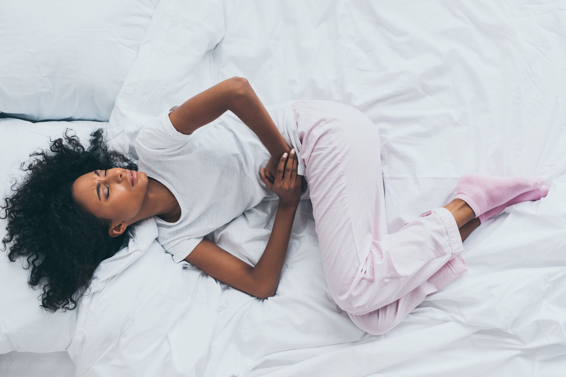 Une personne allongée sur un lit blanc, vêtue d'une chemise blanche et d'un pantalon rose, se tient le ventre