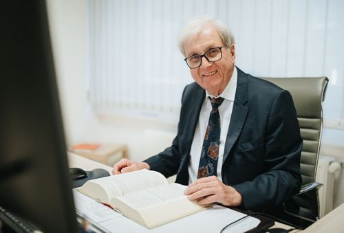 Dr. Rainer Hildebrand in einem Anzug und Krawatte sitzt an einem Schreibtisch und liest ein Buch.