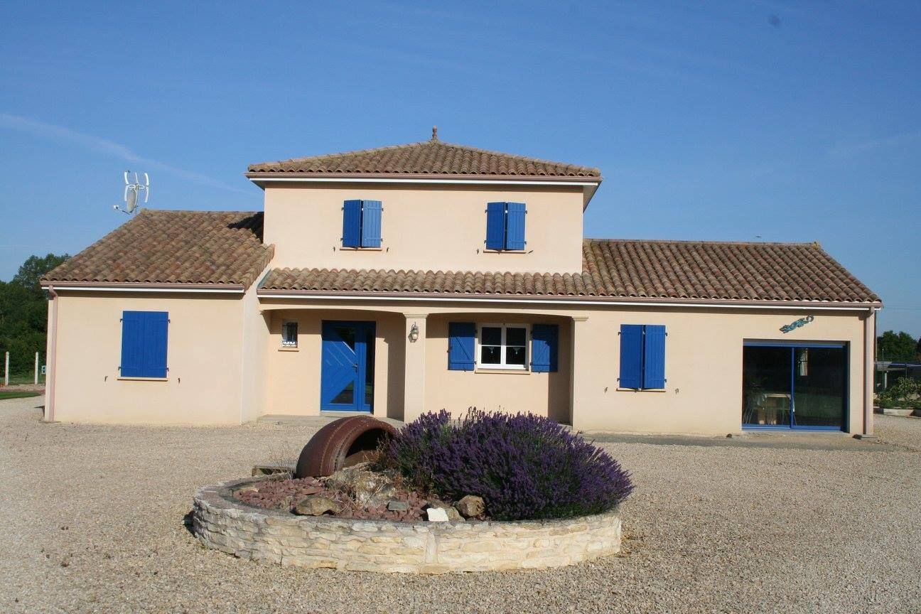 Petite maison beige aux volets et portes bleus, toit de tuiles et cour gravillonnée sous un ciel dégagé.