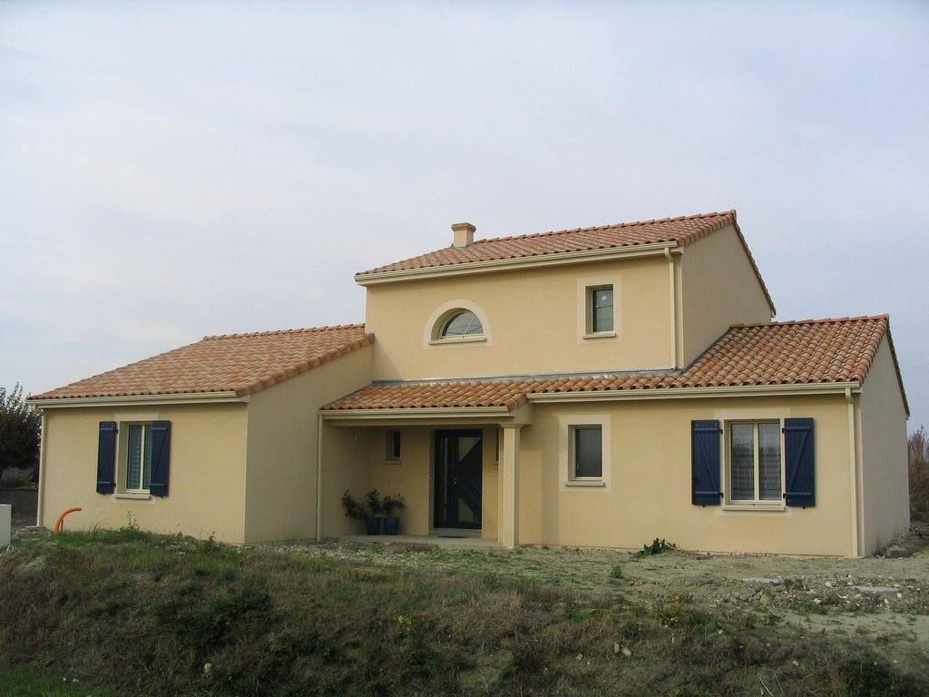 Maison en stuc beige avec toit de tuiles rouges et volets bleus dans une cour herbeuse sous un ciel nuageux.