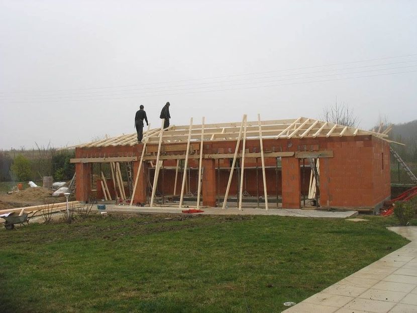Maison en briques en construction, avec deux ouvriers sur la charpente du toit et un premier plan herbeux.