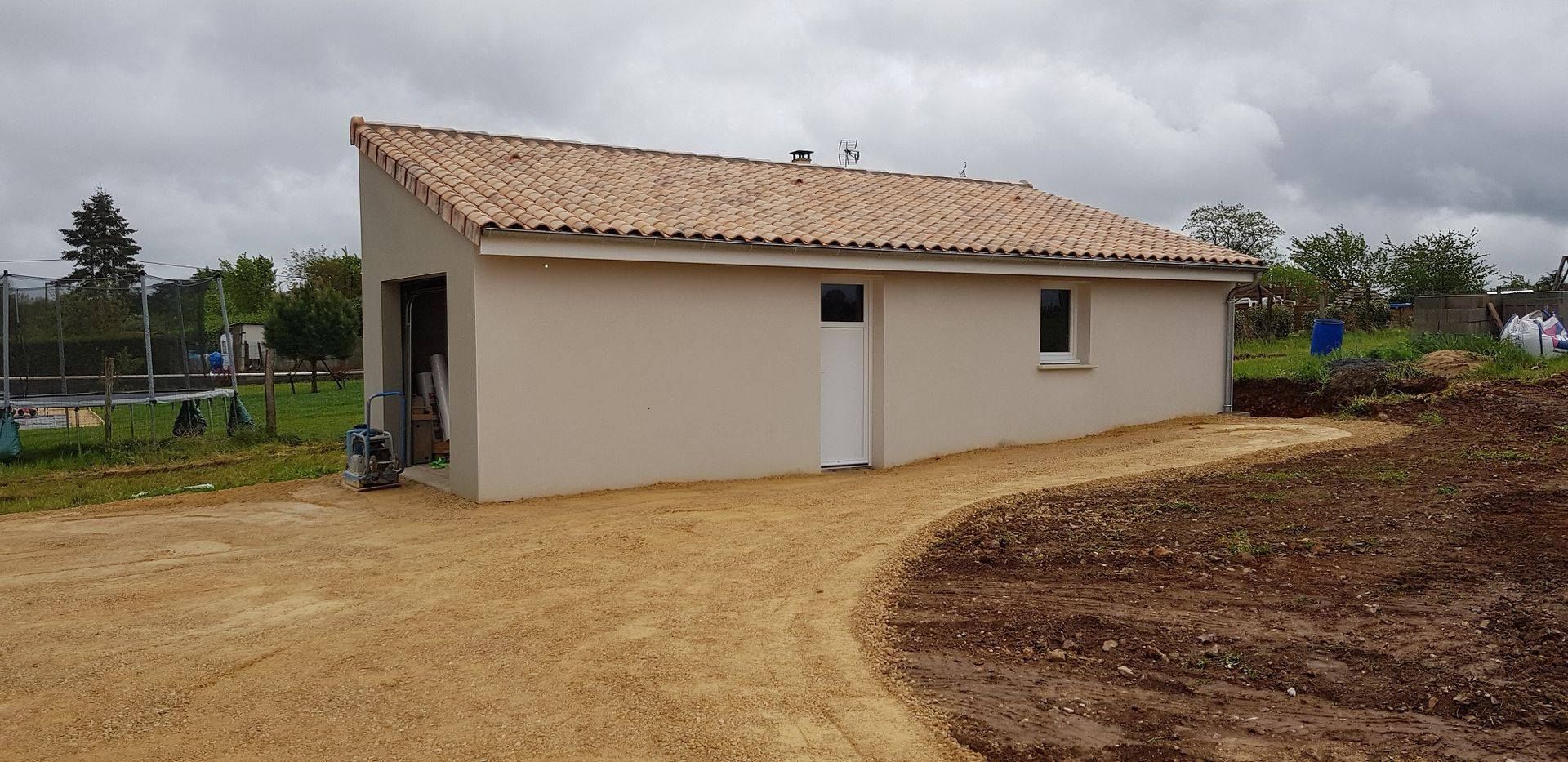 Petite maison beige au toit de tuiles, située sur un chemin de terre sous un ciel nuageux.
