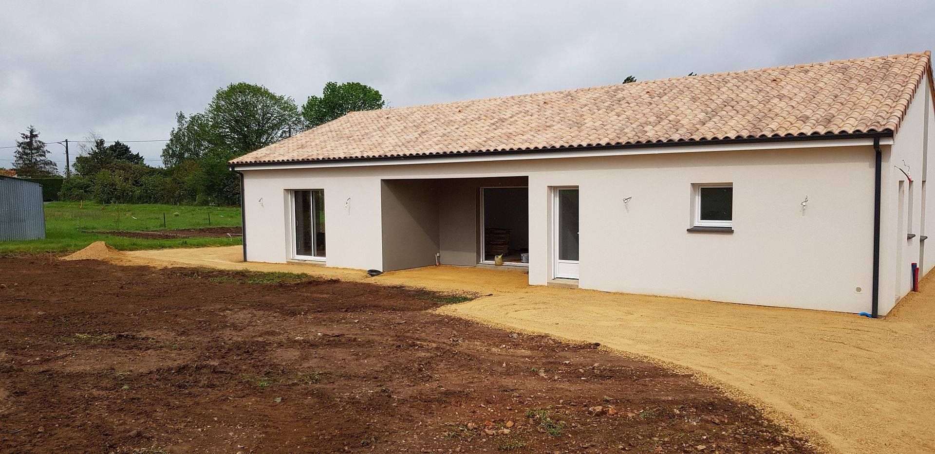 Maison blanche de plain-pied en construction sur un terrain en terre battue, avec un toit de tuiles et une entrée ouverte.