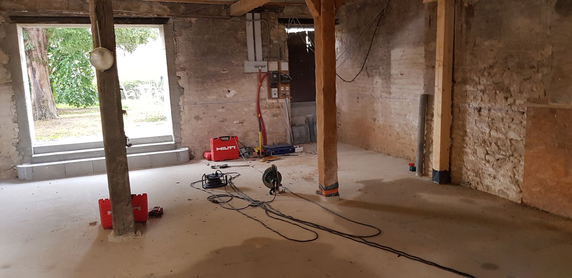 Pièce inachevée avec sol en béton, murs en briques apparentes, poteaux en bois et outils éparpillés près d'une fenêtre.