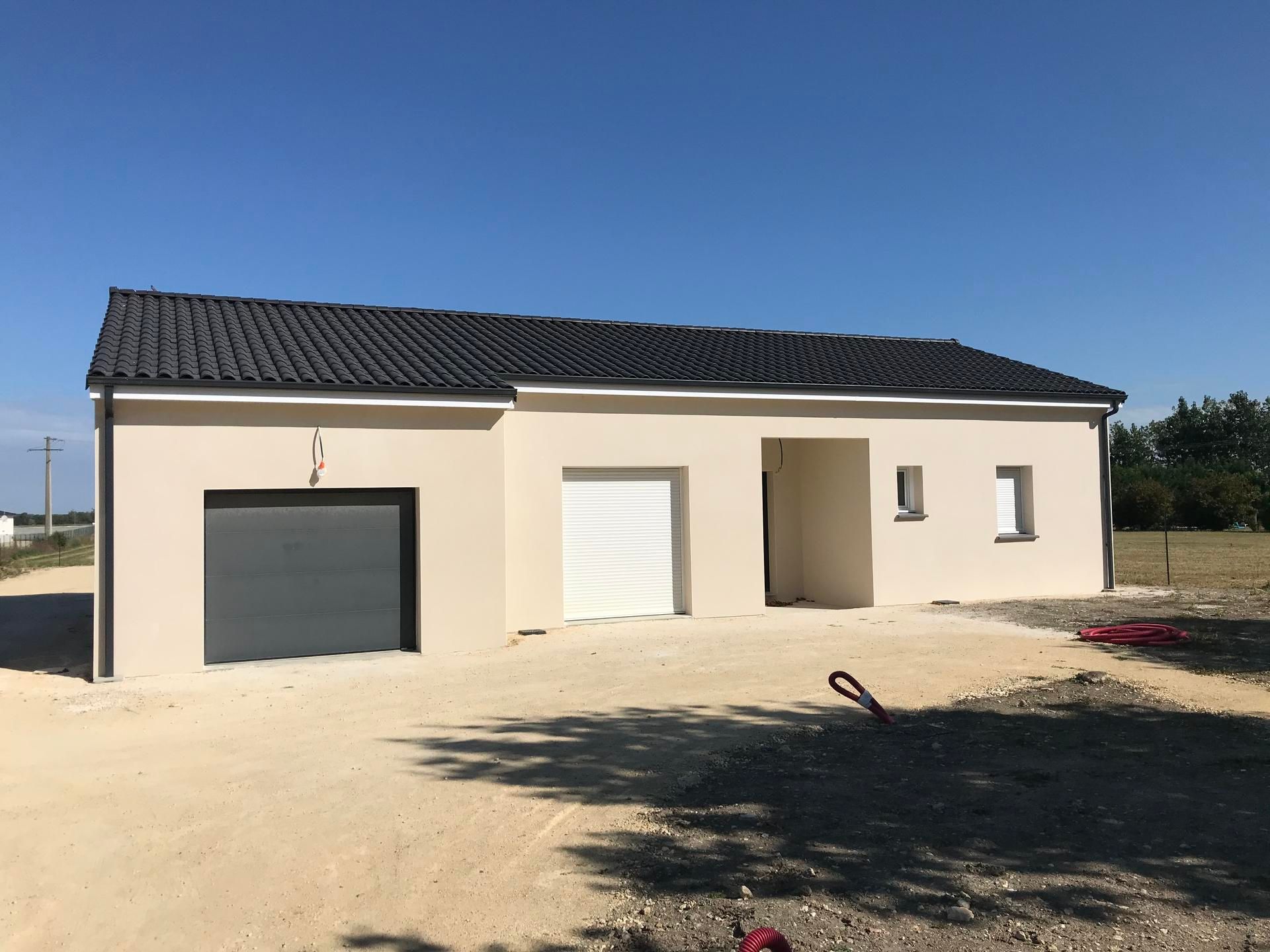 Maison moderne de plain-pied beige avec toit foncé, porte de garage et cour sablonneuse.
