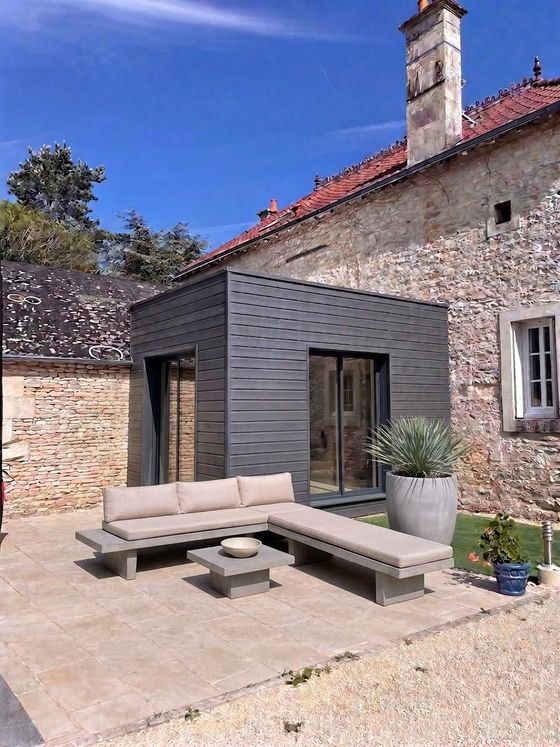 Cour en pierre avec annexe moderne noire et sièges extérieurs beiges adossés à une maison en pierre rustique.