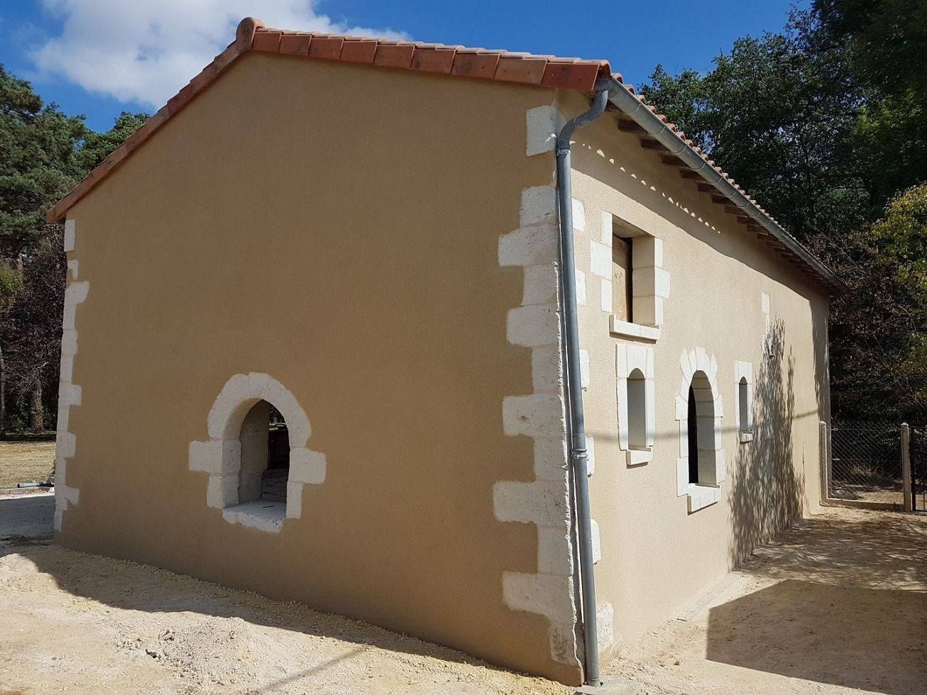 Petite maison en stuc beige avec un toit de tuiles rouges et des fenêtres cintrées dans un cadre rural.