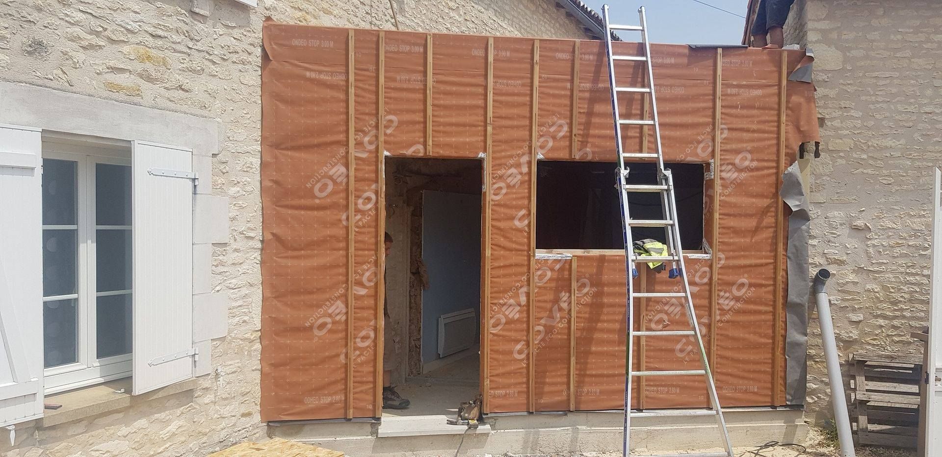 Façade d'un bâtiment en construction, revêtue de panneaux bruns, avec une échelle appuyée contre elle.