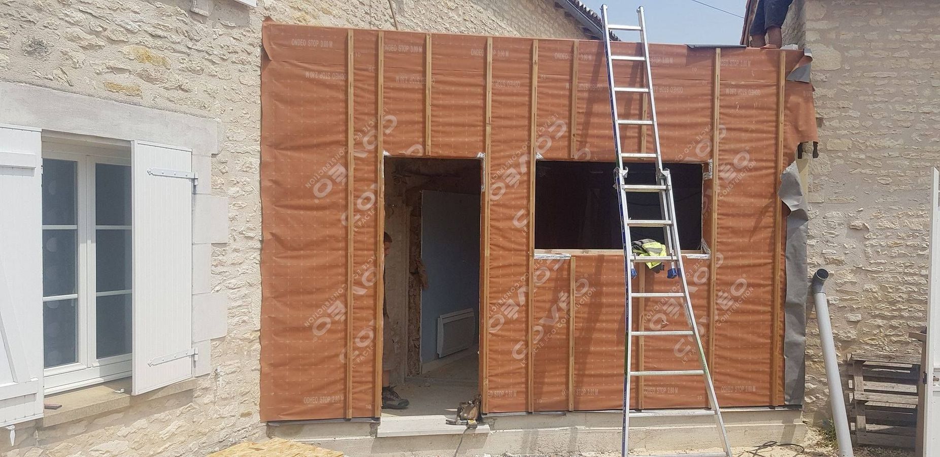 Façade d'un bâtiment en construction, revêtue d'orange, avec une échelle appuyée contre elle et une porte ouverte en dessous.