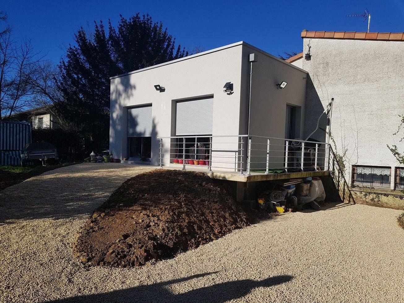 Petite maison blanche avec une rampe d'accès, une allée de gravier et un monticule de terre devant.