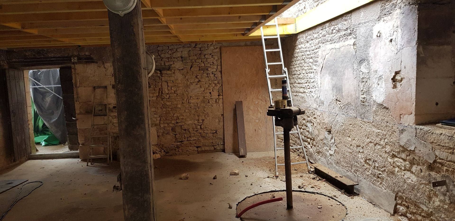 Sous-sol avec murs en pierre apparente, poutres de plafond en bois, échelle, poteaux de soutien et sol en terre battue.