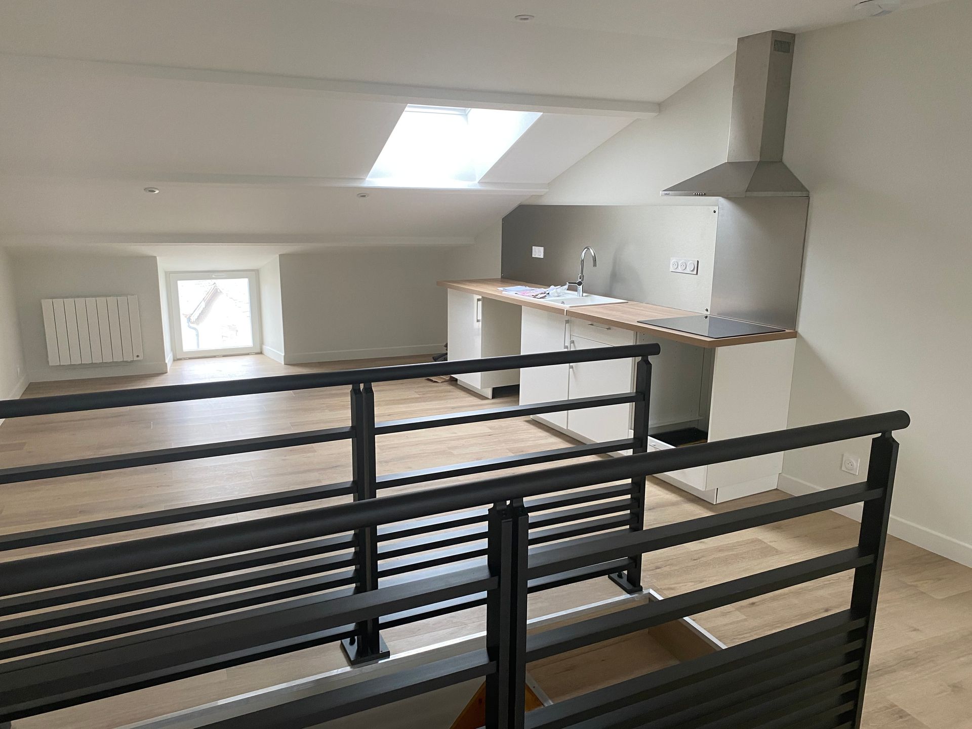 Cuisine loft moderne avec rambardes noires, murs blancs, puits de lumière et armoires en bois clair.