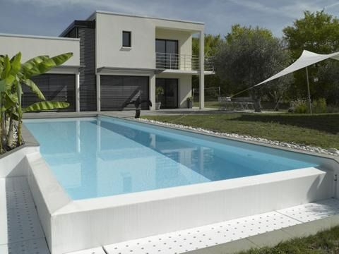 Maison moderne au bord d'une piscine rectangulaire bleue, entourée de pelouse et d'arbres.