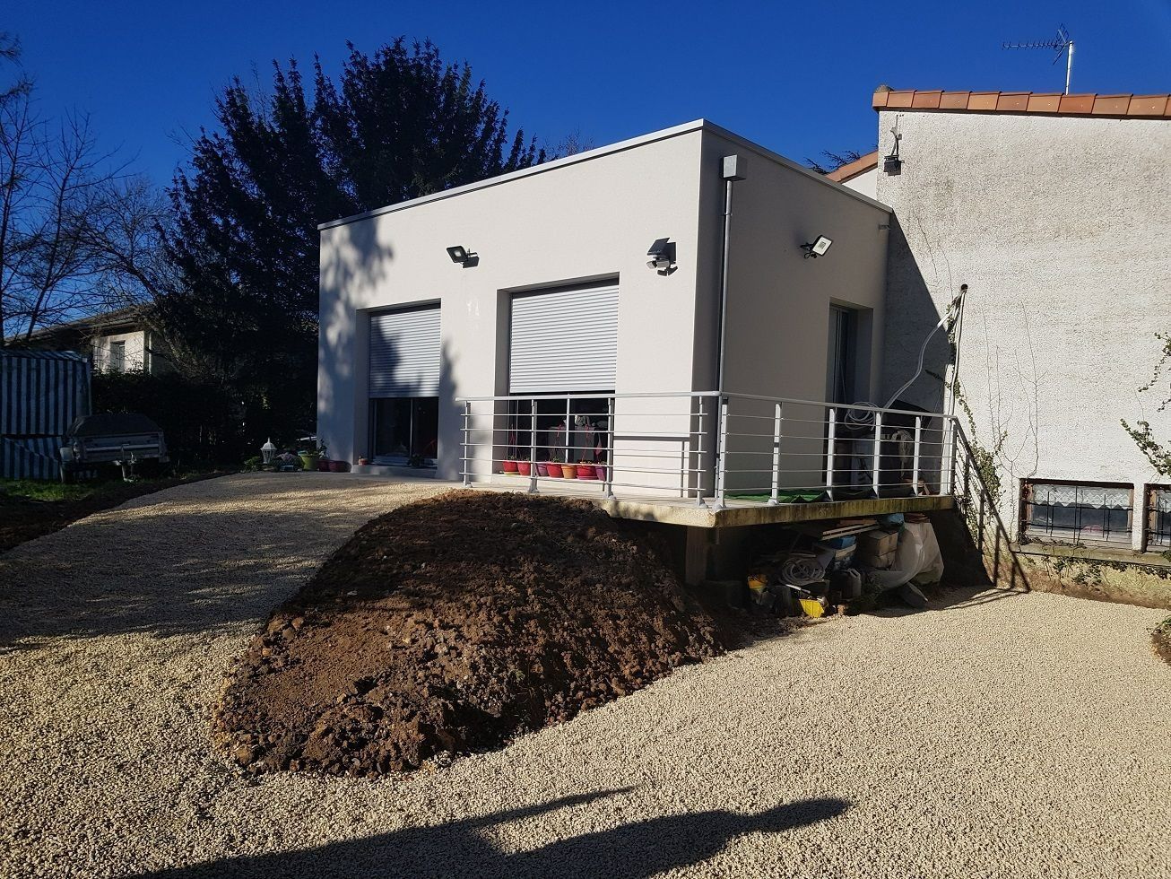 Petite maison blanche avec une terrasse surélevée, une cour en gravier et un grand tas de terre devant.