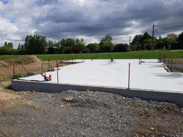 Dalle de fondation en béton en construction sur un terrain gravillonné, avec des piquets et un ciel nuageux.