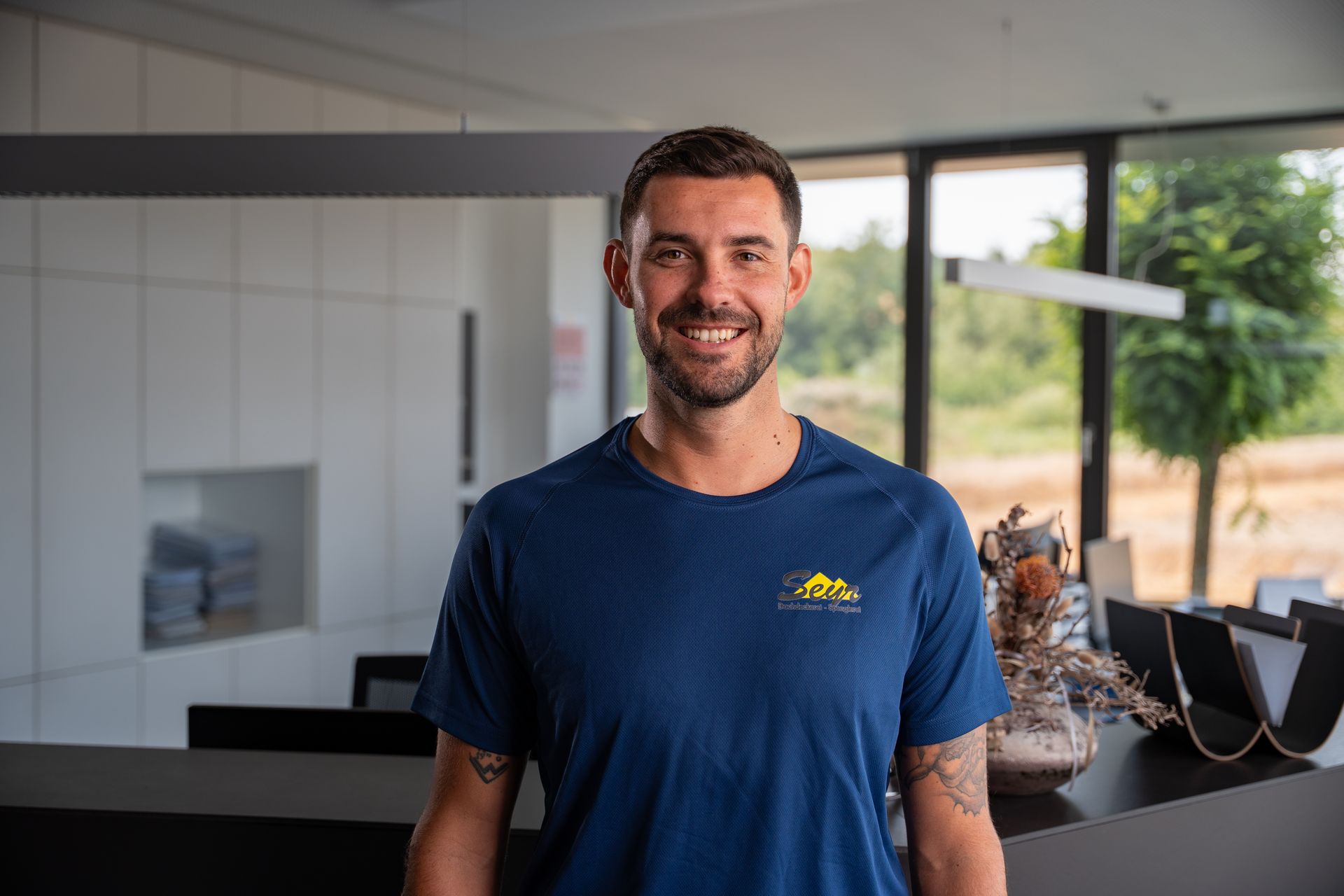 Ein Mann mit Bart trägt ein blaues Hemd und steht mit verschränkten Armen da.