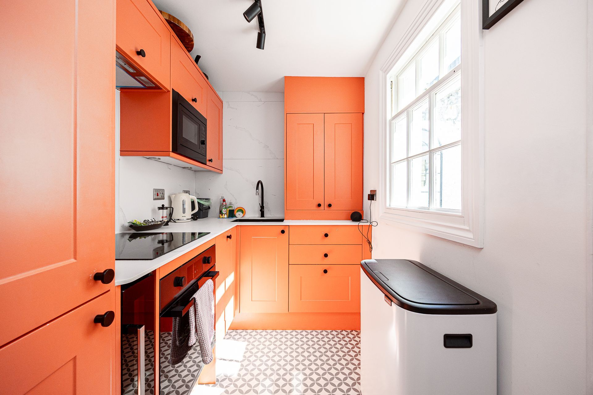 Bright orange galley kitchen with white countertop, patterned floor, and a window by a small trash bin