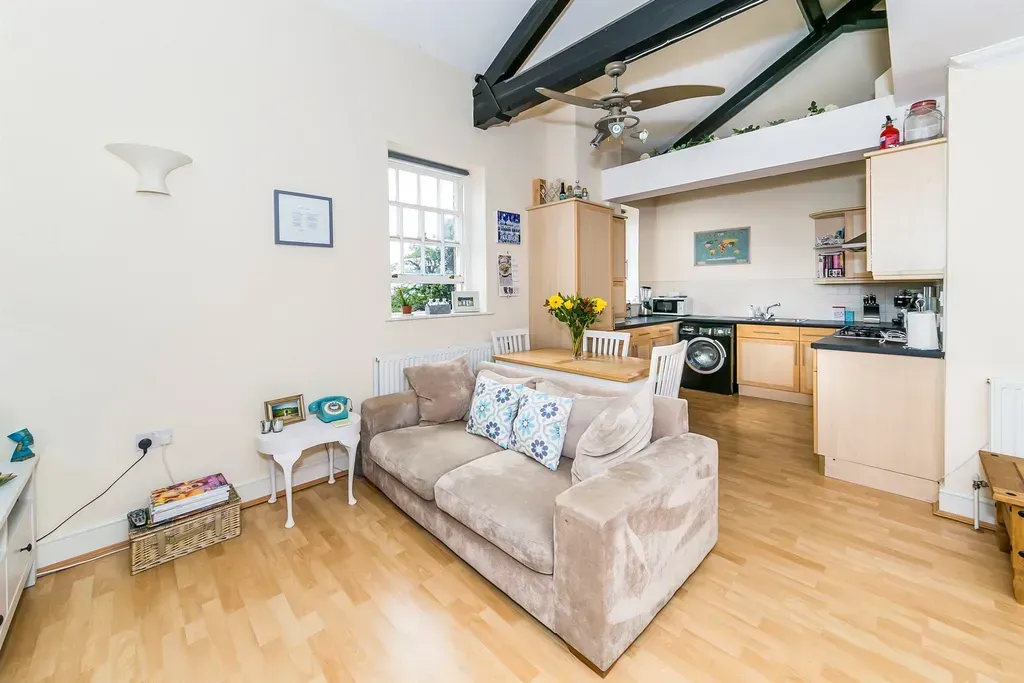 Bright open living room with beige sofa, wood floors, exposed beams, and kitchen area in back.