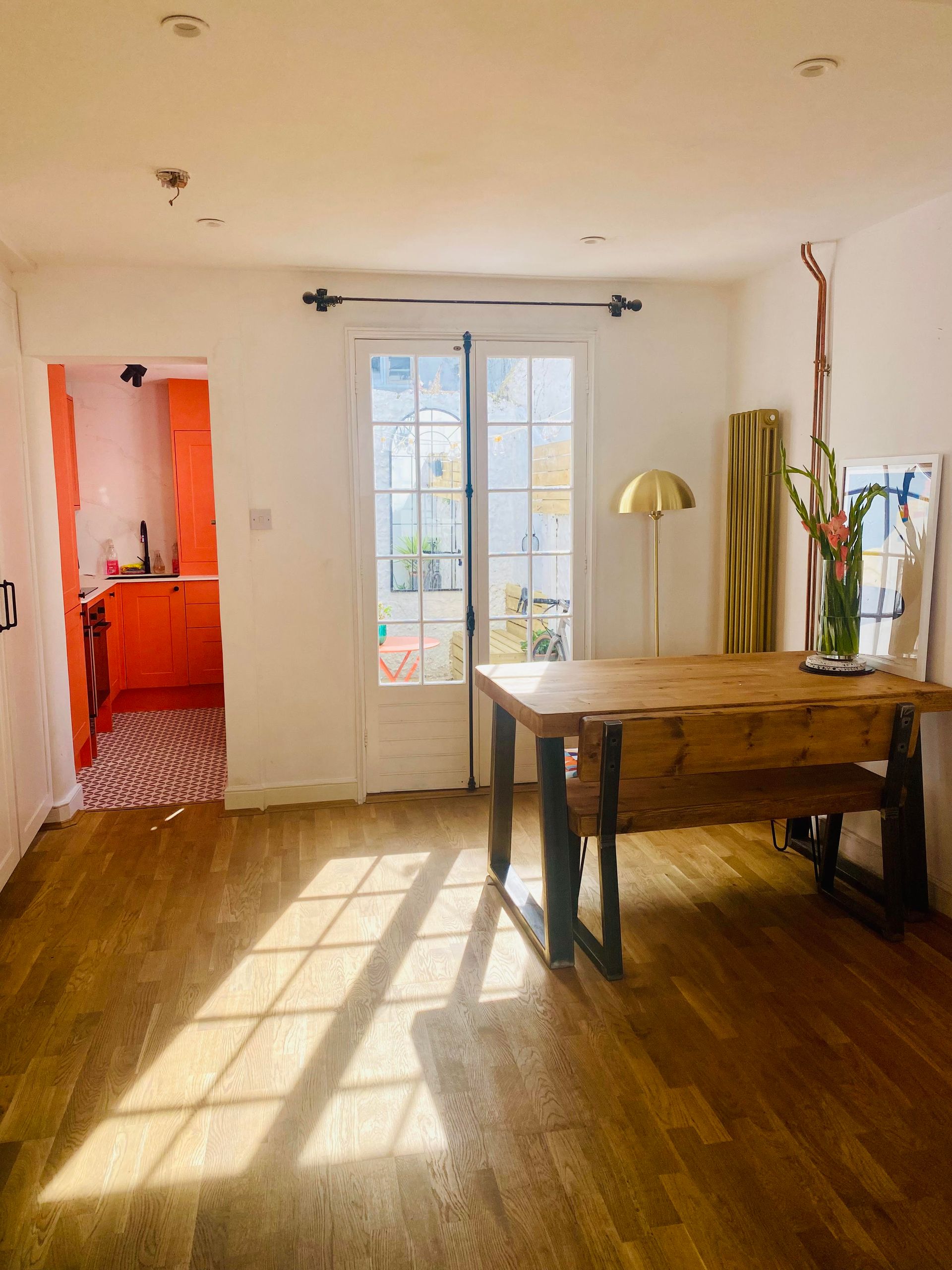 Bright living room with wooden floor, sunlit French doors, table, lamp, and orange kitchen doorway.