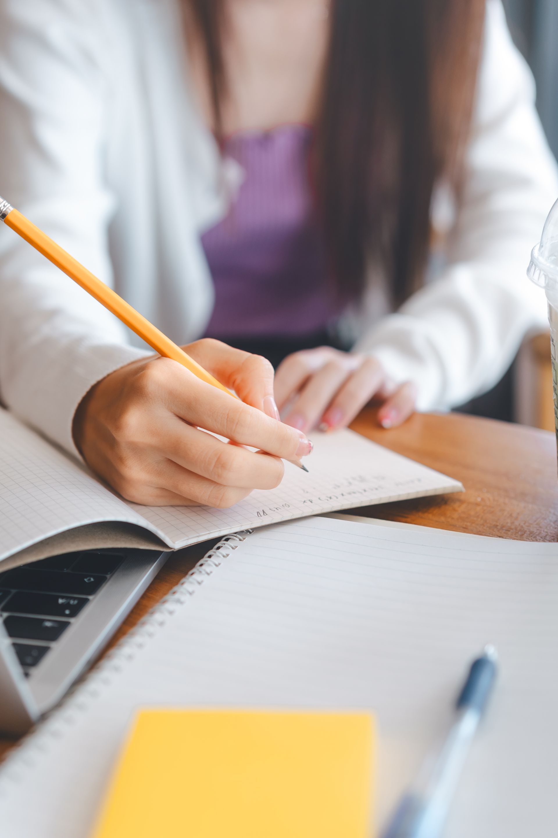 Femme écrivant dans un cahier avec un crayon, un ordinateur portable, des notes autocollantes et un stylo sur une table en bois.