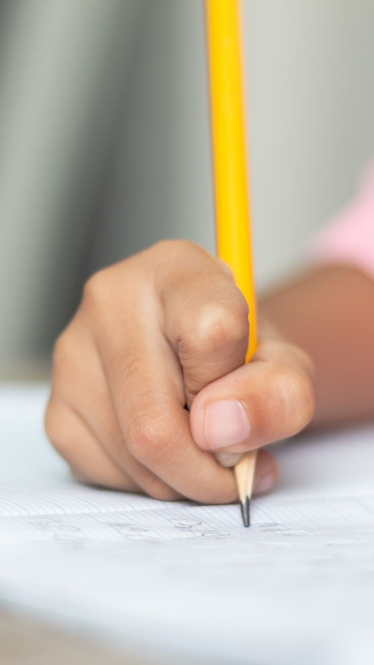 Main d'enfant tenant un crayon jaune et écrivant sur une feuille blanche. Gros plan, éclairage tamisé.