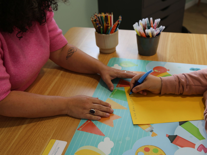 Femme âgée souriante aux cheveux gris dans une chemise jaune, écrivant à une table dans une cuisine.