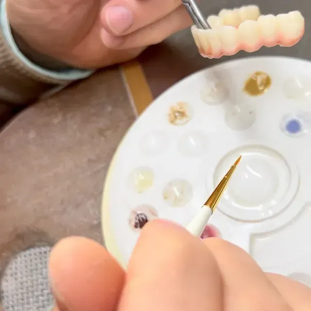Persona pintando detalles de prótesis dentales con un pincel, utilizando una paleta de colores.