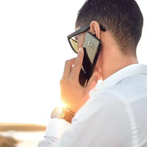 Hombre con camisa blanca y gafas de sol, sosteniendo un teléfono en su oreja, al aire libre.