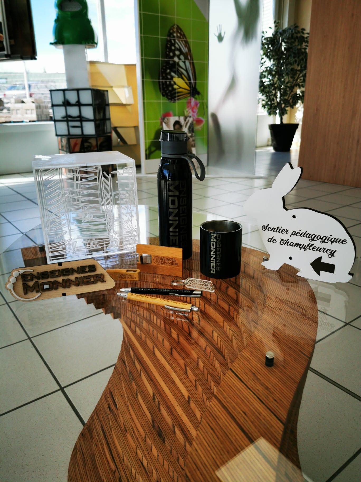 Petite table d'exposition avec un assortiment de bouteilles, de tasses et une pancarte découpée, dans un espace intérieur carrelé.