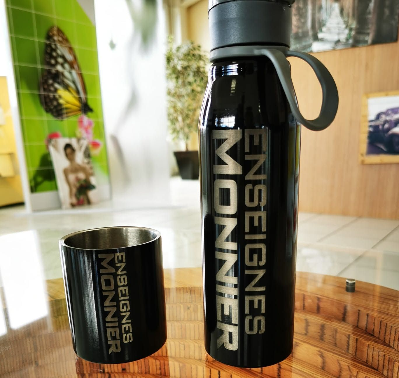 Bouteille isotherme noire et tasse sur une table en bois, toutes deux marquées au nom de l'entreprise.