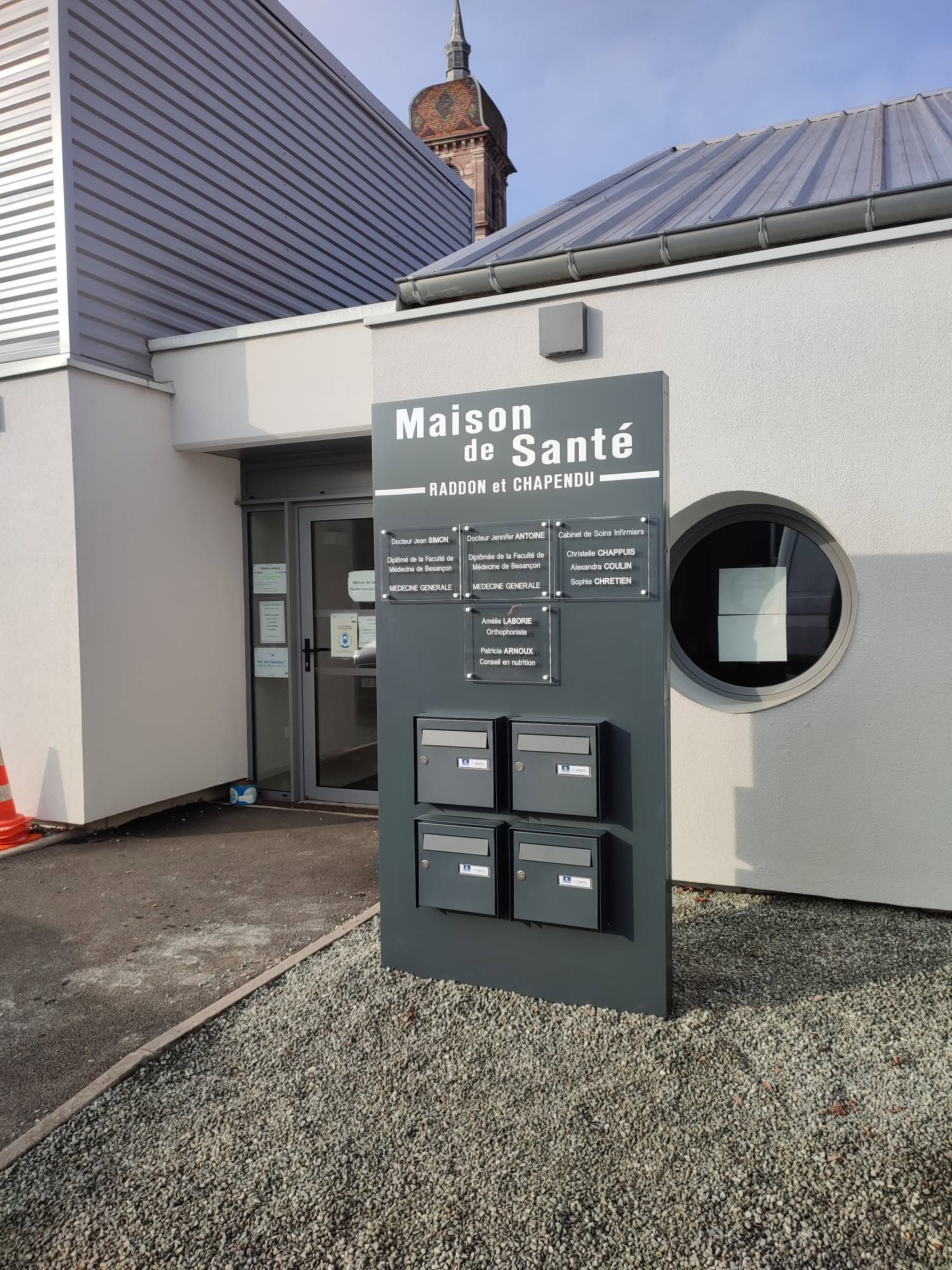 Panneau d'entrée Maison de Santé devant une clinique moderne, avec une façade grise et une fenêtre ronde.