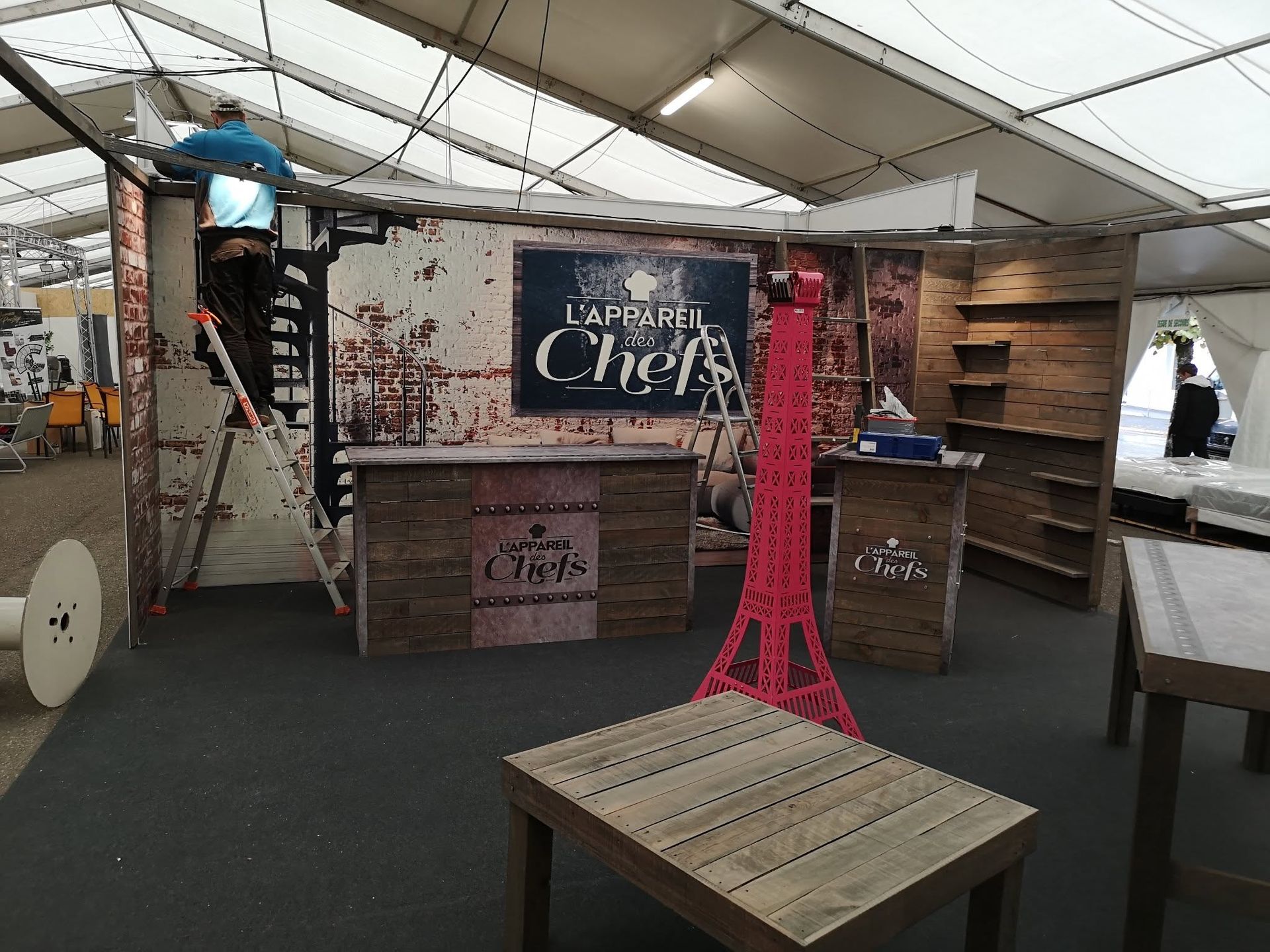 Étal de marché avec des caisses en bois, une ardoise et une décoration rouge représentant la Tour Eiffel sous un toit en toile de tente.