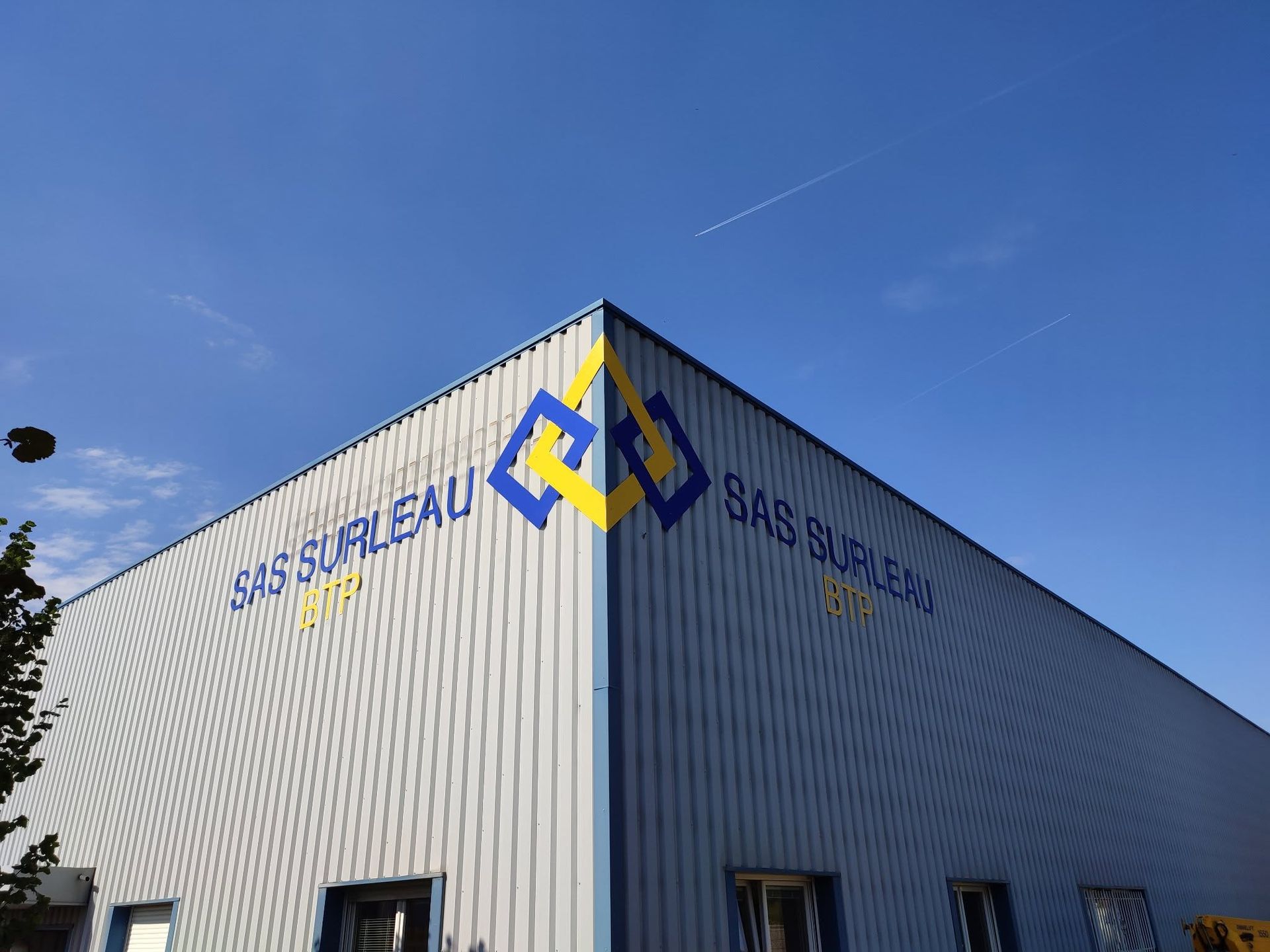 Logo bleu et jaune sur un bâtiment industriel en tôle ondulée sous un ciel bleu dégagé.