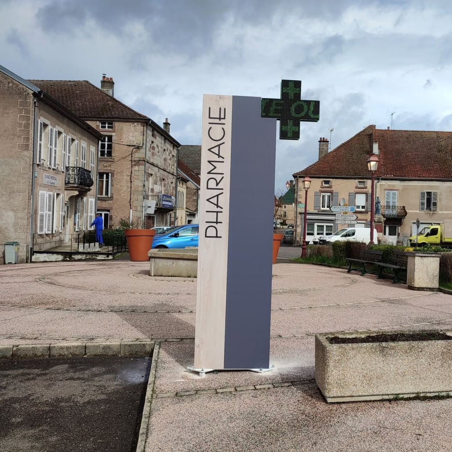 Enseigne de pharmacie extérieure avec une croix verte sur une place de village.