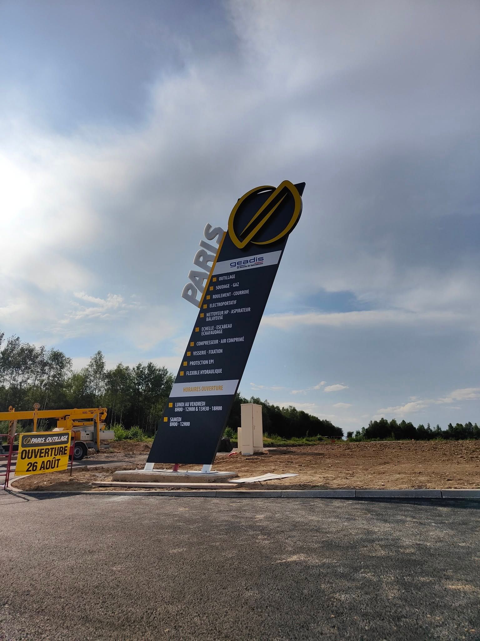 Grand panneau routier noir avec des accents jaunes sur un terrain gravillonné sous un ciel nuageux.