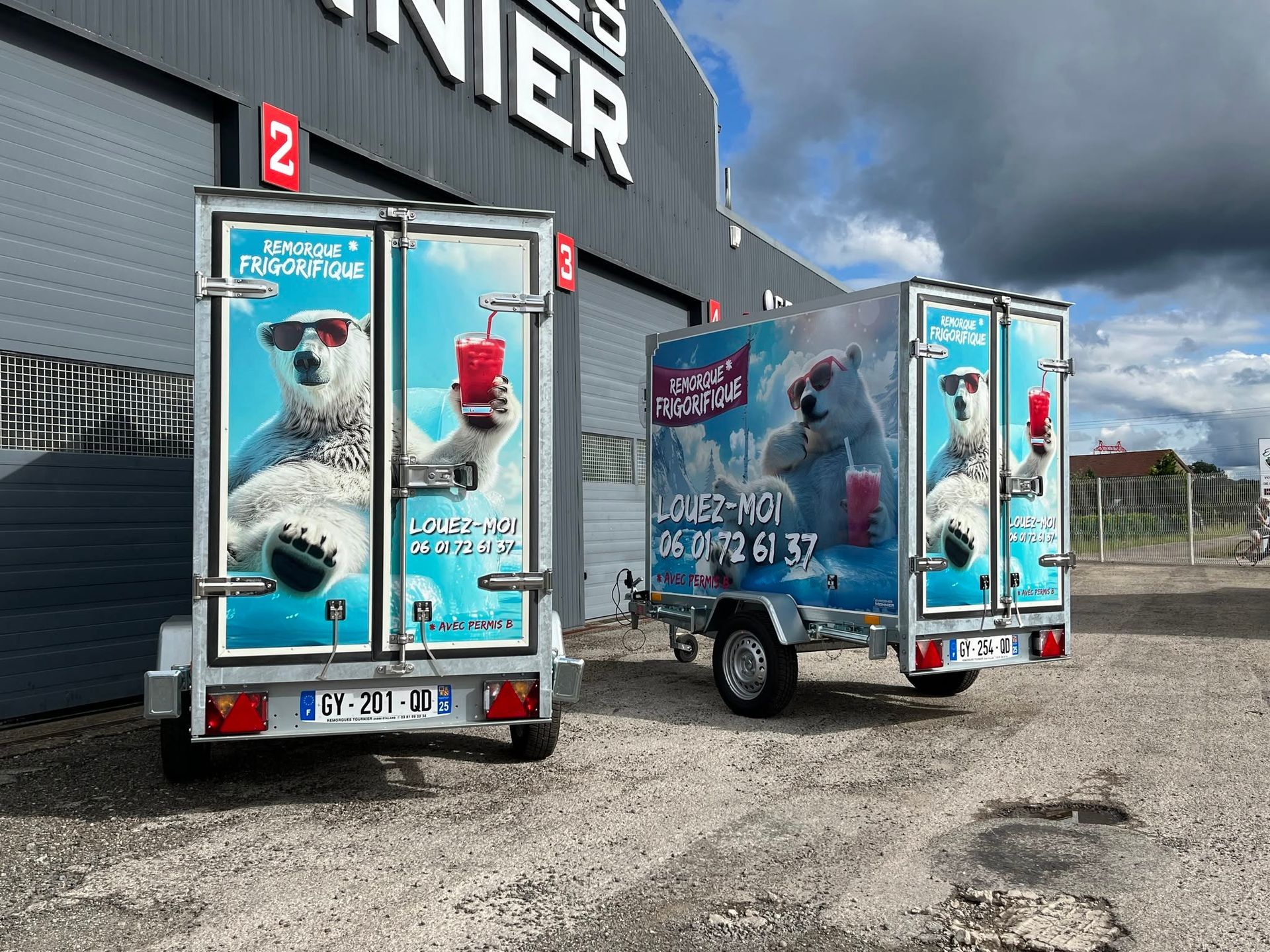 Deux remorques bleues de vente ambulante, ornées de publicités sont stationnées devant un bâtiment sous un ciel nuageux.