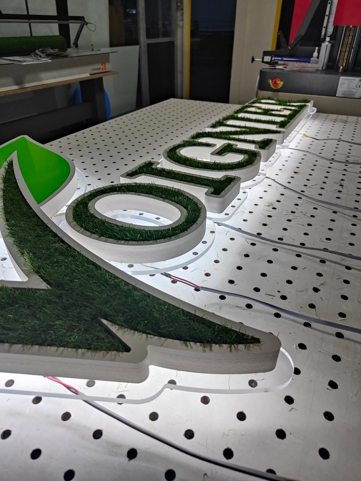 Des lettres en mousse verte sur une table de travail perforée dans un atelier, formant une partie d'une enseigne.