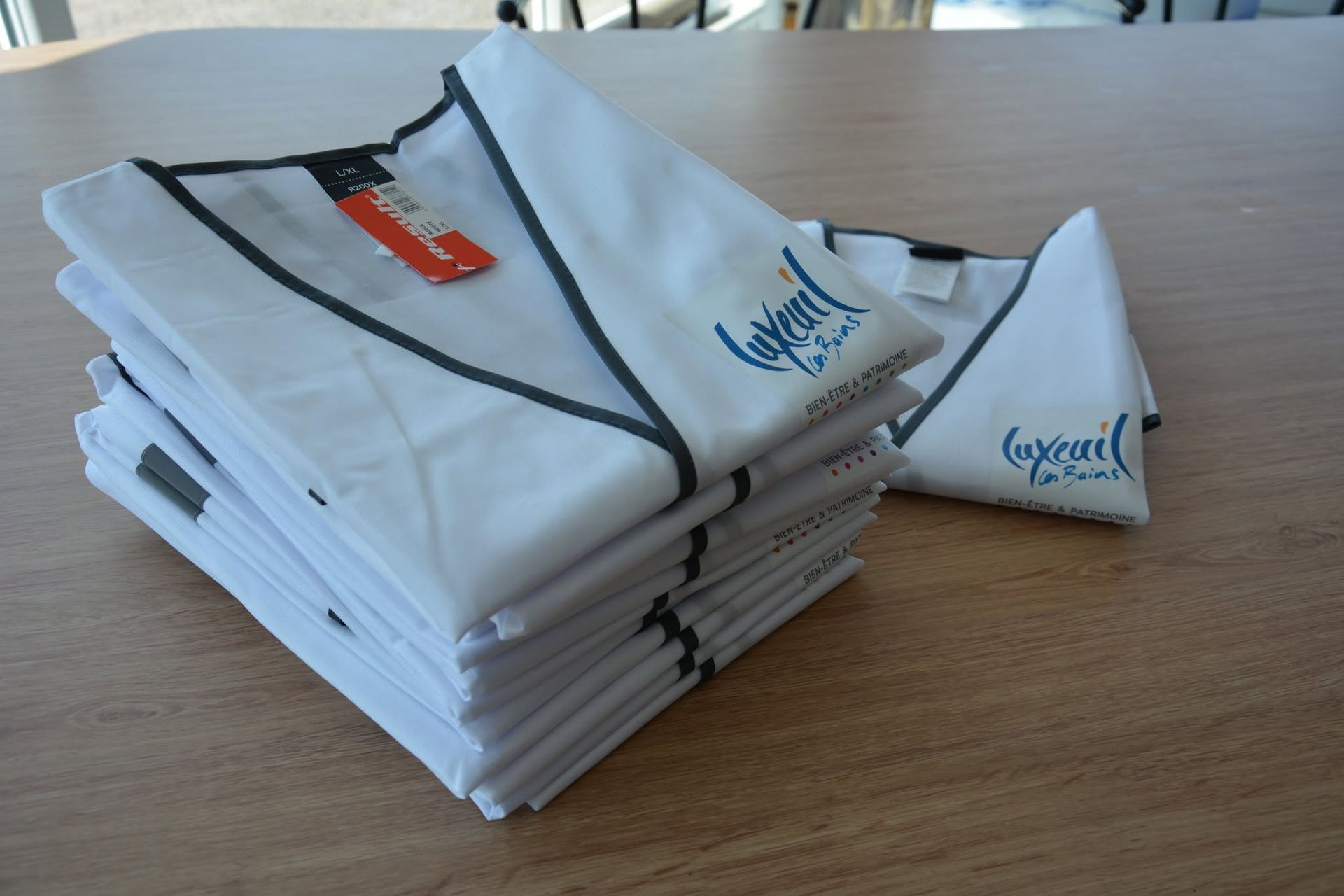 Pile de blouses blanches pliées, bordées de liseré foncé et ornées d'un logo bleu.