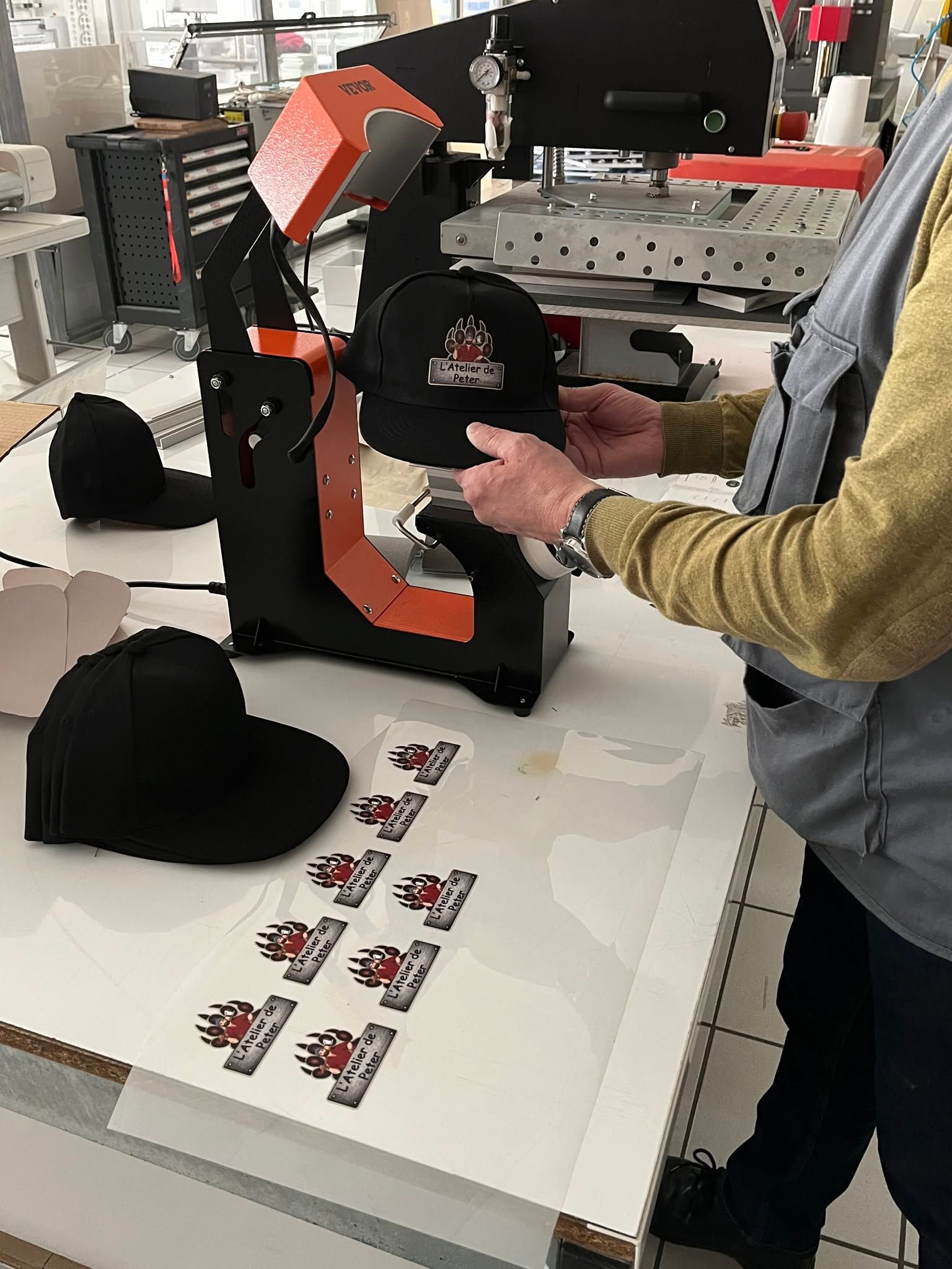 Une personne ajuste une presse à chaud noire et orange à côté de chapeaux et d'articles imprimés sur une table de travail.