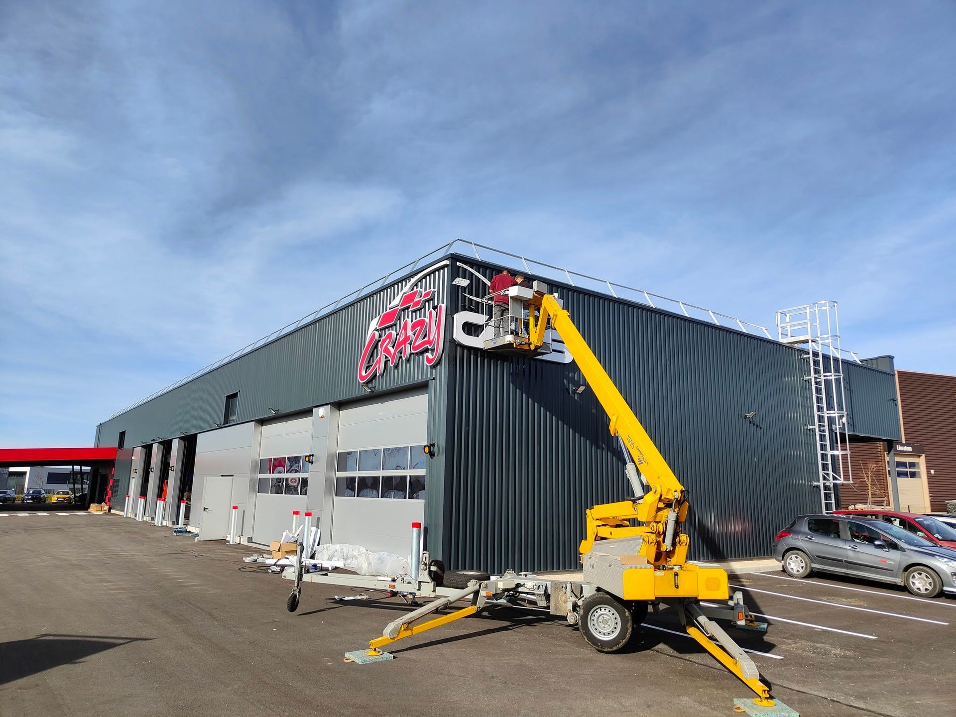 Une nacelle jaune en fonctionnement à l'extérieur d'un bâtiment commercial gris avec une signalétique rouge, sous un ciel bleu.