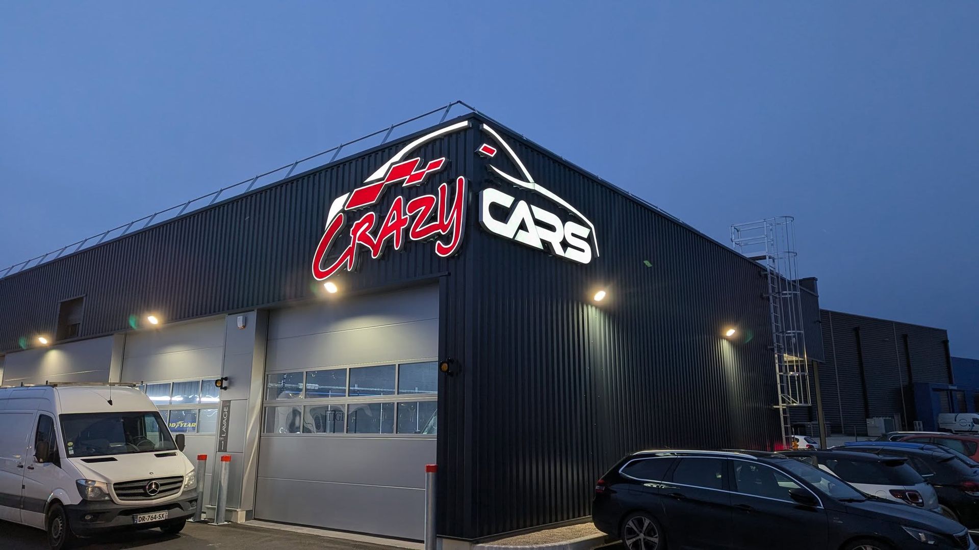 Façade d'une entreprise au crépuscule, avec extérieur noir, enseigne lumineuse et véhicules stationnés.