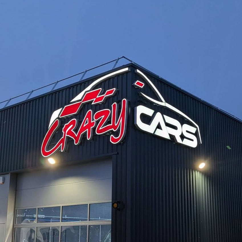 Enseigne du concessionnaire Crazy Cars sur un bâtiment sombre au crépuscule, avec des lettres rouges et blanches sur fond de ciel bleu.